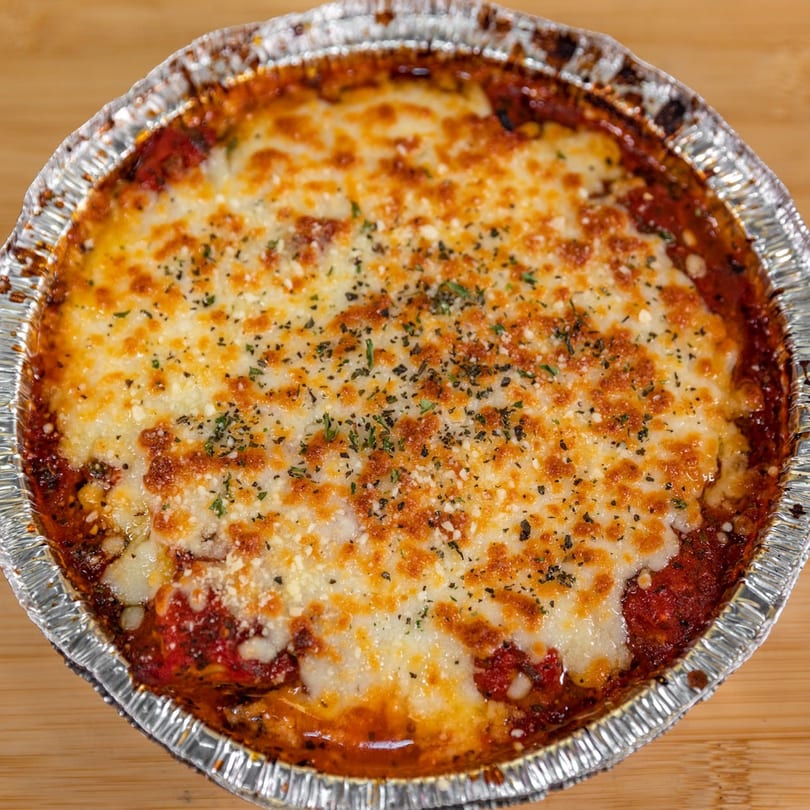 Baked pasta dish from La Pizzeria, topped with melted cheese and herbs in a foil container.